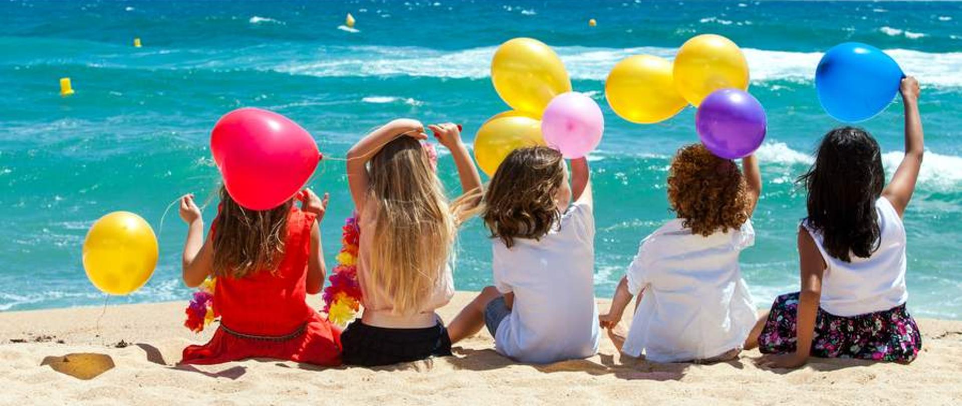 Małe dzieci trzymające kolorowe balony siedzące na plaży.
