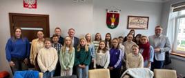 Grupa dzieci i młodzieży wraz z opiekunkami oraz pracownikami Urzędu Miasta na tle jasnej ściany, na której wisi godło państwowe, herb miasta Lubawa oraz stara panorama miasta.