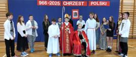 Na środku na tronie siedzi uczeń przebrany za króla Bolesława Chrobrego, obok stoi uczeń w przebraniu biskupa z pastorałem i uczeń przebrany za pomocnika księdza w białej szacie, za nimi w półokręgu stoją pozostali uczniowie klasy 7 odgrywający role społeczeństwa