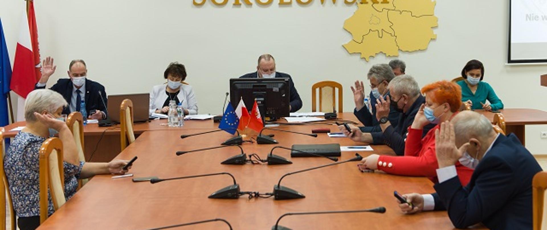 Na zdjęciu siedzący przy stole w Sali konferencyjnej Starostwa Powiatowego w Sokołowie Podlaskim radni Rady Powiatu Sokołowskiego głosują uchwałę poprzez podniesienie ręki. Na ścianie widnieje złoty napis Powiat Sokołowski oraz przestrzenna mapa powiatu z podziałem na poszczególne gminy i miasta, a także symbol orła w koronie oraz katolicki krzyż z Jezusem Chrystusem.