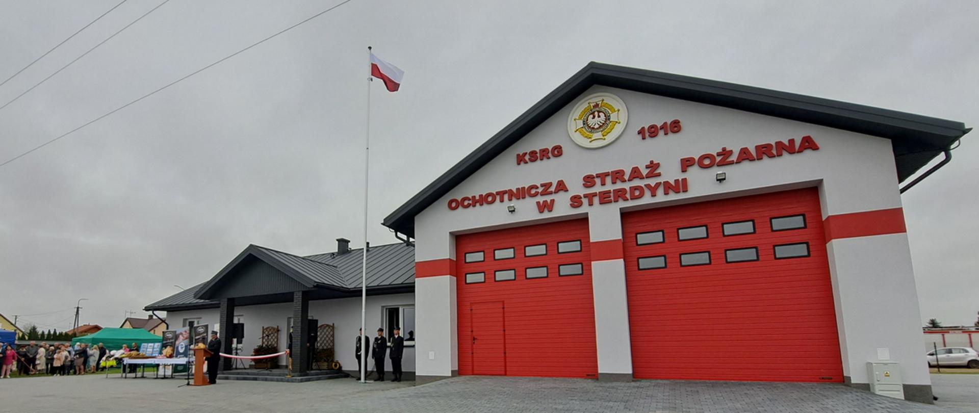 Na zdjęciu widzimy nową siedzibę OSP w Sterdyni. Na pierwszym planie na placu wyłożonym szarą kostką brukową widzimy budynek z dwuspadowym dachem o szarej elewacji przedzielonej czerwonym poprzecznym pasem z parą dużych czerwonych rozsuwanych drzwi (bramy wjazdowe do hangaru dla dwóch wozów strażackich). Na elewacji ponad drzwiami widzimy czerwone napisy KSRG logo ZOSP RP 1916, a pod spodem Ochotnicza Straż Pożarna w Sterdyni. Po lewej stronie od hangaru poprzeczna dwuspadowa przybudówka z gankiem opartym na kolumnach, w której znajdują się pomieszczenia socjalne i baza strażacka. Po środku widzimy maszt z powiewającą flagą Polski. Wokół widzimy również uczestników uroczystości otwarcia remizy.