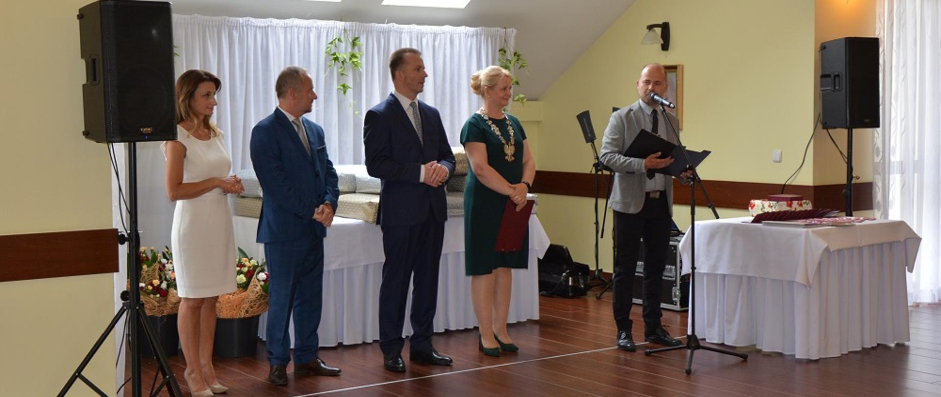 dwie kobiety i trzech mężczyzn odświętnie ubranych, witają zebranych gości 