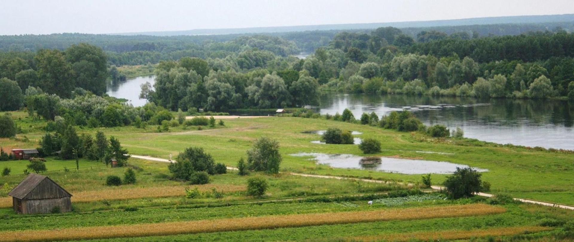 Krajobraz z widokiem na zielone łąki i rzekę z dużą ilością drzew, w lewym dolnym rogu stodoła. Zdjęcie przedstawia tereny województwa podlaskiego.