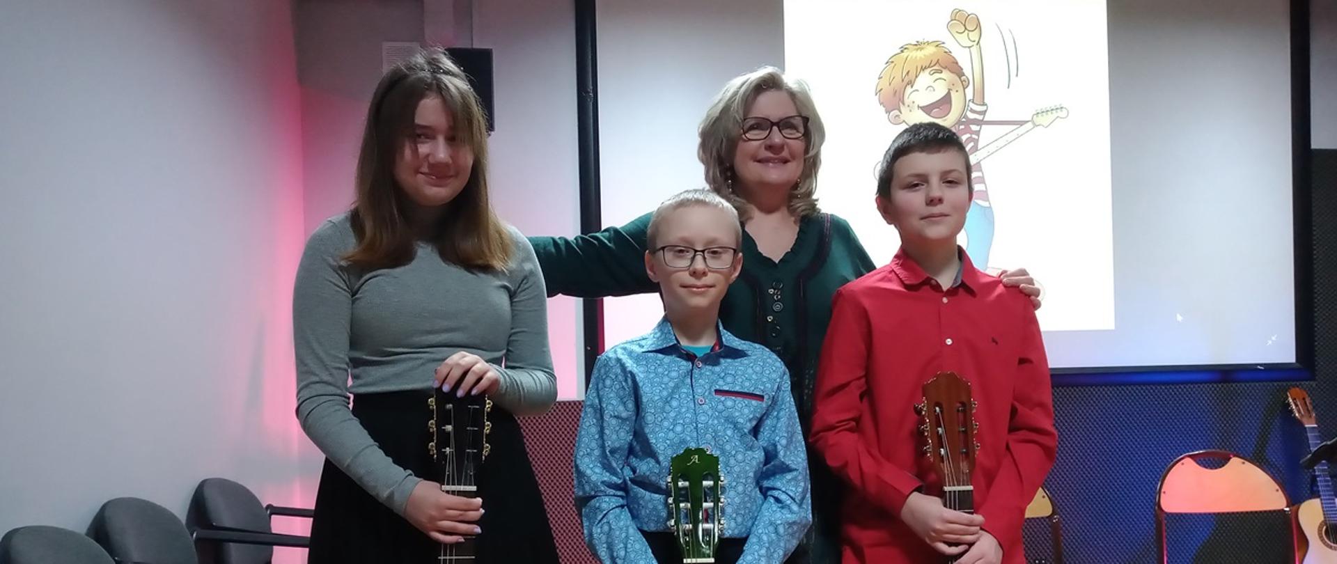 Troje dzieci stoi i przed sobą trzyma gitary. Za nimi stoi kobieta