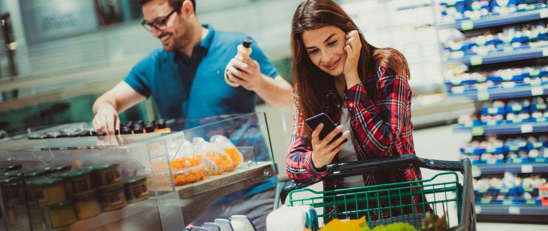 Na zdjęciu widać kobietę i mężczyznę robiących zakupy w supermarkecie. Mężczyzna wybiera napój, kobieta stoi przy wózku i trzyma w ręce telefon