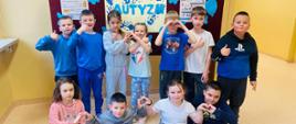 Uczniowie stoją przed gazetką informacyjną, na której są zawieszone informacje na temat autyzmu oraz niebieskie motylki i baloniki, pokazują z dłoni symbol serduszka
