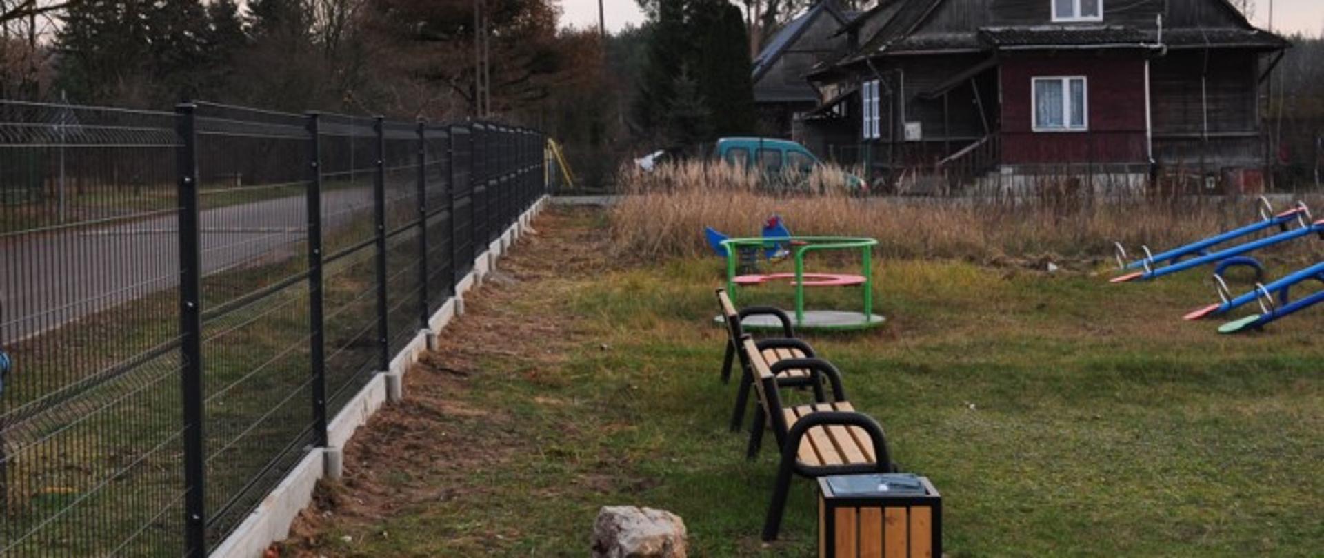 Na zdjęciu widoczne nowe ogrodzenie placu zabaw w miejscowości Dalekie Tartak. Ogrodzenie wykonano z paneli stalowych z cokołem z płyt betonowych. Na zdjęciu znajdują się także umieszczone na placu zabaw ławki, karuzela oraz huśtawki. W tle widać drewniane domy oraz drzewa.