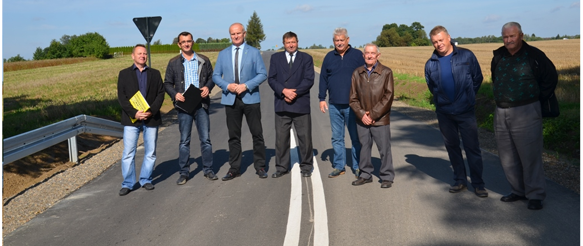 Na zdjęciu widać grupę ludzi stojących w rzędzie na jezdni. W tle widzimy drogę oraz po bokach rozciągają się pola