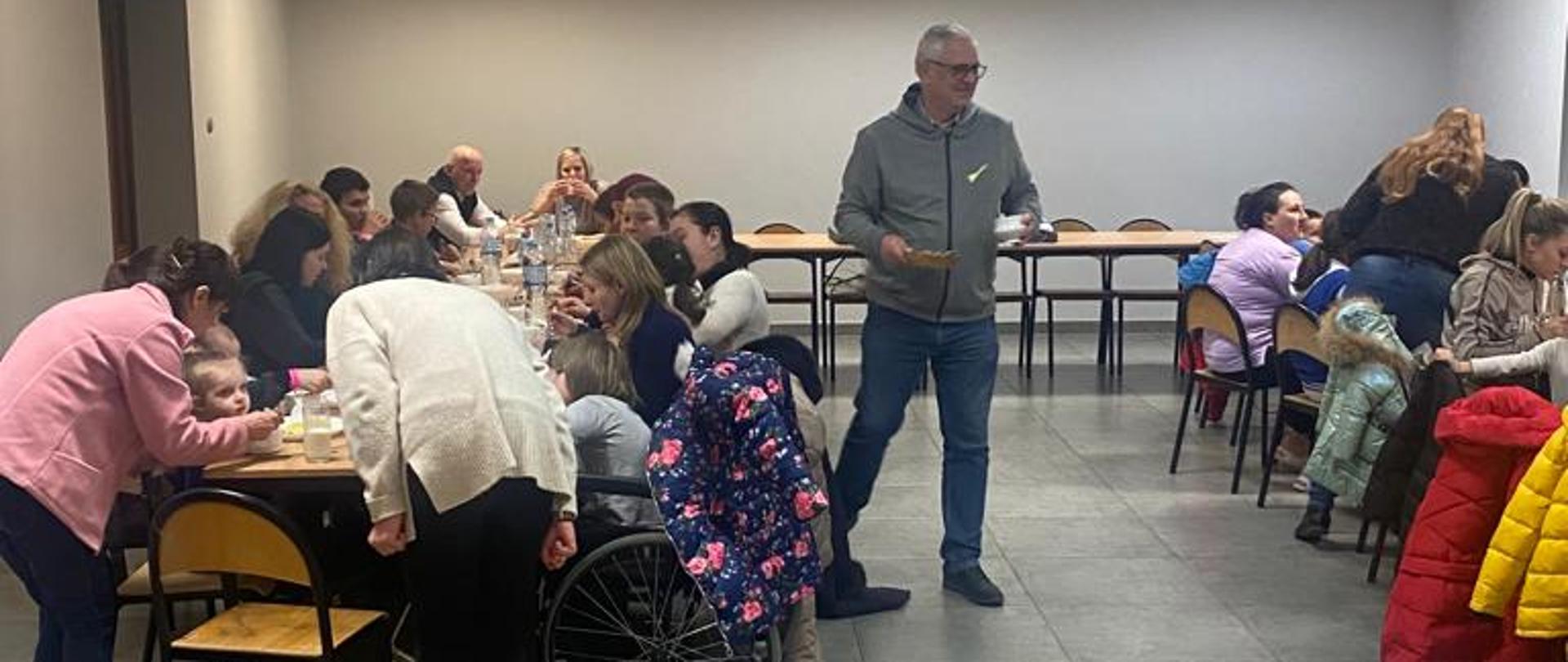 ludzie siedzący na krzesłach przy stołach w sali Miejsko-Gminnego Ośrodka Kultury w Margoninie, trzy osoby stoją, dwie z nich pochylają się nad stołami