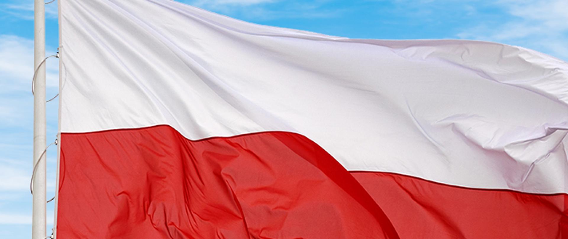 Flaga Narodowa Rzeczypospolitej Polskiej powiewa na maszcie na tle błękitnego nieba.