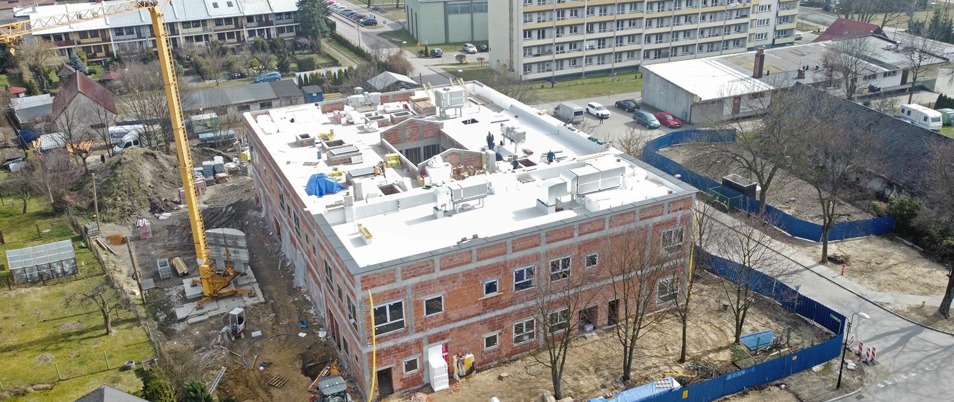 Widok z góry na budowane przedszkole, obok stoi dźwig a w tle zabudowania mieszkalne.
