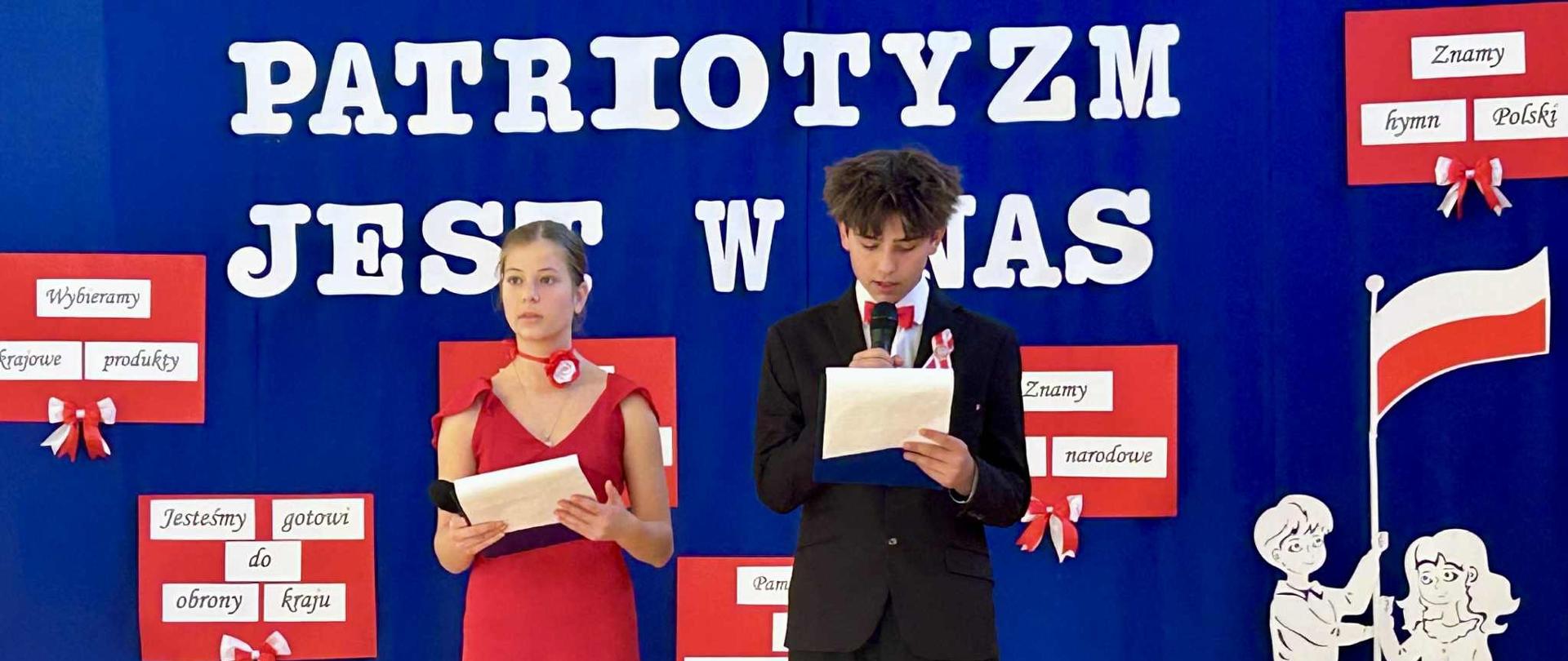 Dwoje dzieci ( chłopiec w czarnym garniturze, białej koszuli i czerwonej muszce oraz dziewczynka w czerwonej sukience) stoi na sali gimnastycznej. W tle dekoracja - na szfirowym płótnie napis z białych liter "Patriotyzm jest w nas".