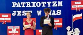 Dwoje dzieci ( chłopiec w czarnym garniturze, białej koszuli i czerwonej muszce oraz dziewczynka w czerwonej sukience) stoi na sali gimnastycznej. W tle dekoracja - na szfirowym płótnie napis z białych liter "Patriotyzm jest w nas".