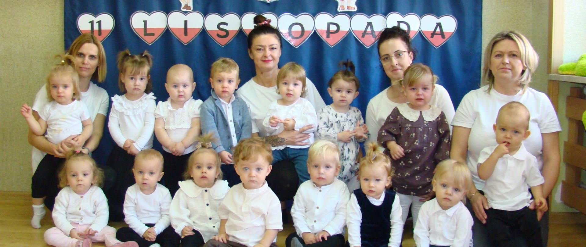 Na zdjęciu są dzieci i panie z grupy Pszczółki.Wszyscy są ubrani nw "galowe" stroje. W tle, na granatowym materiale widnieje napis "11 Listopada" oraz symbole narodowe: godło i flaga.
