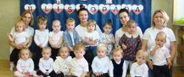 Na zdjęciu są dzieci i panie z grupy Pszczółki.Wszyscy są ubrani nw "galowe" stroje. W tle, na granatowym materiale widnieje napis "11 Listopada" oraz symbole narodowe: godło i flaga.