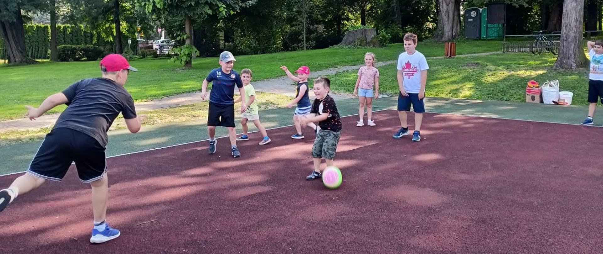 Dzieci grają w piłkę na boisku. Jedno dziecko kopie kolorową piłkę, a reszta obserwuje lub uczestniczy w zabawie. W tle rosną wysokie drzewa i znajduje się ścieżka parkowa.