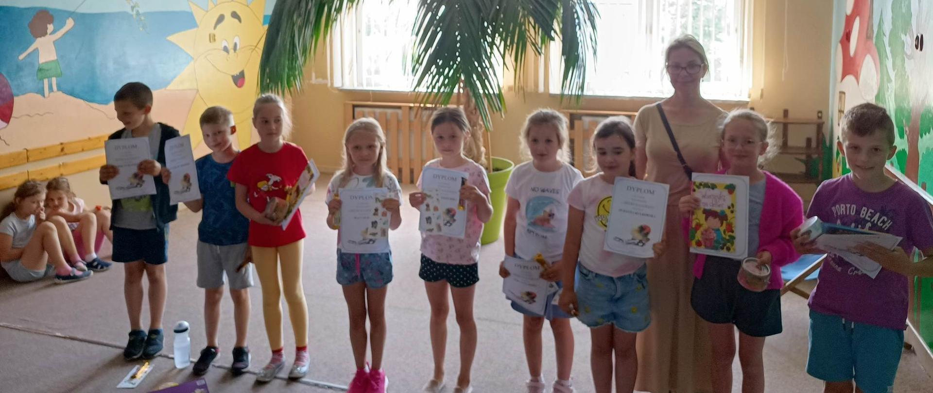 9 dzieci stoi i trzyma przed sobą kolorowe książki, za nimi stoi kobieta