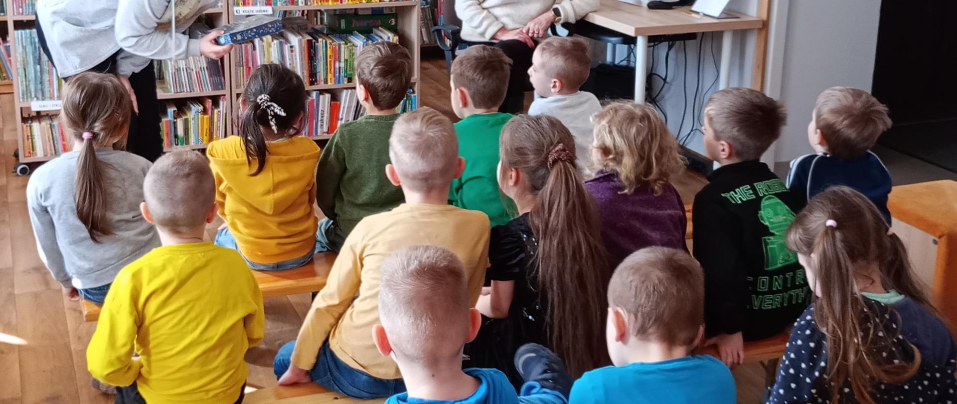 Na zdjęciu widać dzieci przedszkolne siedzące na ławkach i oglądające prezentowaną przez bibliotekarkę książkę