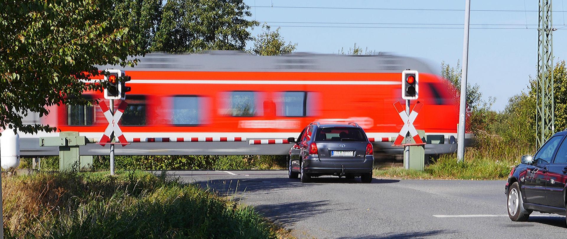 Dwa samochody stojące przed opuszczonym szlabanem przejazdu kolejowego przez który przejeżdża lokomotywa.