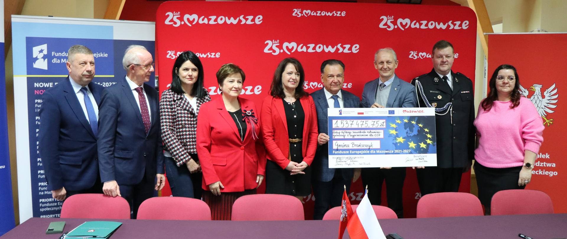 5 mężczyzn i 4 kobiety stojące na tle czerwonej ścianki z napisem 25. lat Mazowsze. Trzech mężczyzn trzyma symboliczny czek.