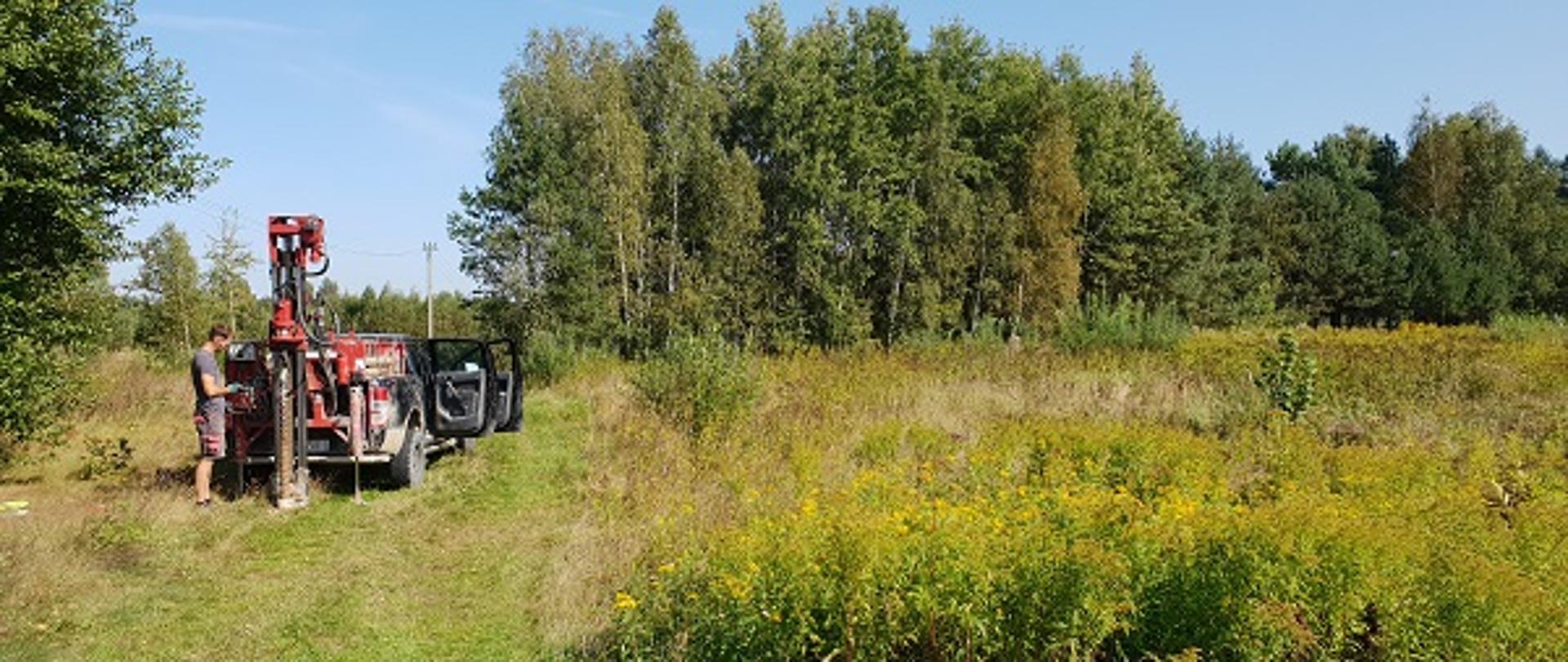 Na zdjęciu znajduje się porośnięta trawami działka w oddali młody lasek. W lewej części fotografii znajduje się samochód osobowy wraz ze specjalistycznym sprzętem przy pomocy którego osoba na zdjęciu wykonuje prace geotechniczne. 