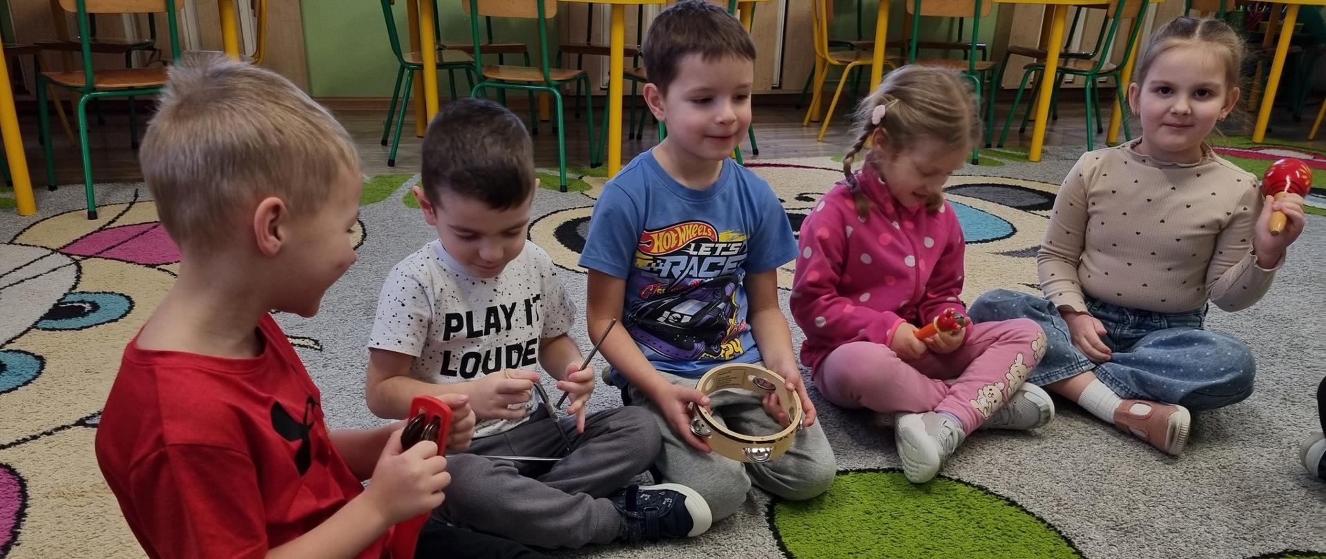 Grupa dzieci siedzi na dywanie grając na instrumentach