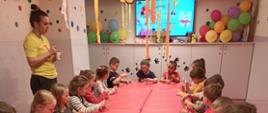 Dzieci siedzące przy długim czerwonym stole. Nad nimi wiszą balony i wstążki