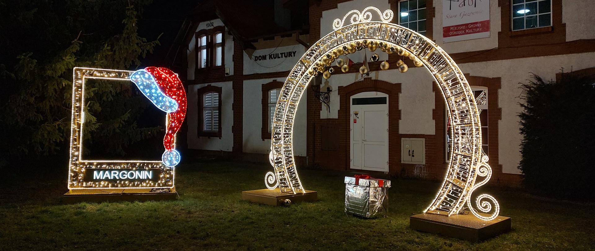 brama iluminacje, w niej prezent owinięty w srebrną folię. po lewej fotobudka z czapką mikołaja i napisem Margonin.