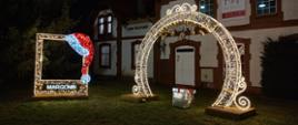 brama iluminacje, w niej prezent owinięty w srebrną folię. po lewej fotobudka z czapką mikołaja i napisem Margonin.