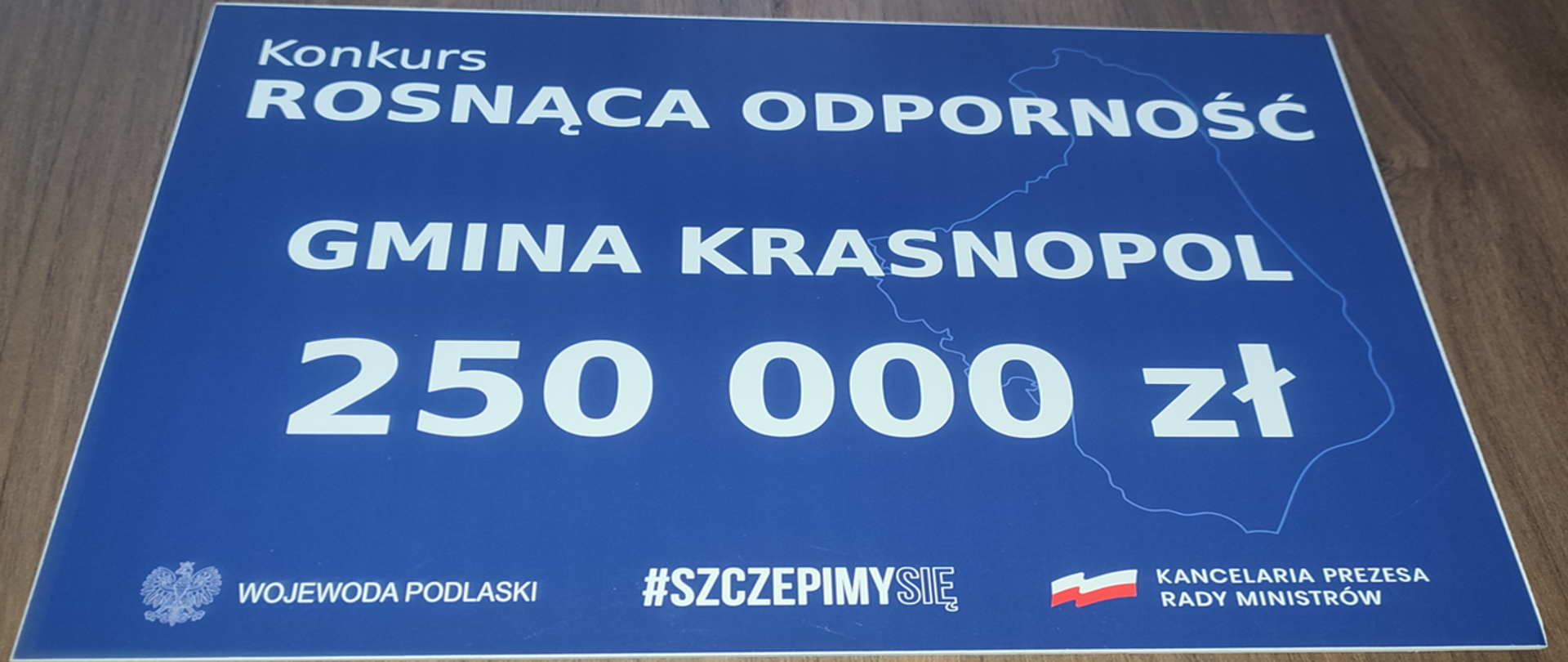 Zdjęcie przedstawia tabliczkę w kolorze granatowym, czcionka koloru białego, widnieją na niej napisy : konkurs Rosnąca odporność Gmina Krasnopol 250 000,00 zł na dole tabliczki są loga: wojewody podlaskiego, szczepimy się oraz logo Kancelarii Prezesa Rady Ministrów
