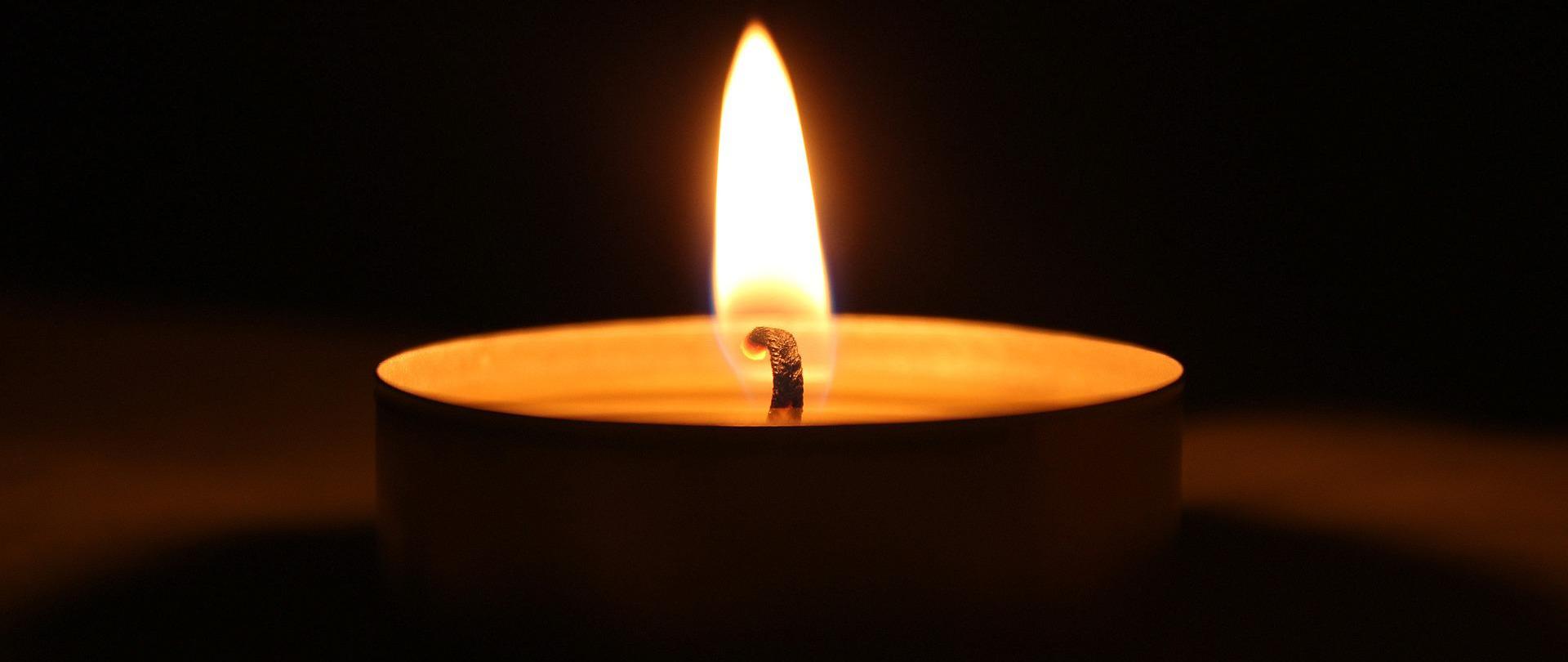 zdjęcie przedstawia na ciemnym tle płonącą świeczkę typu tealight.