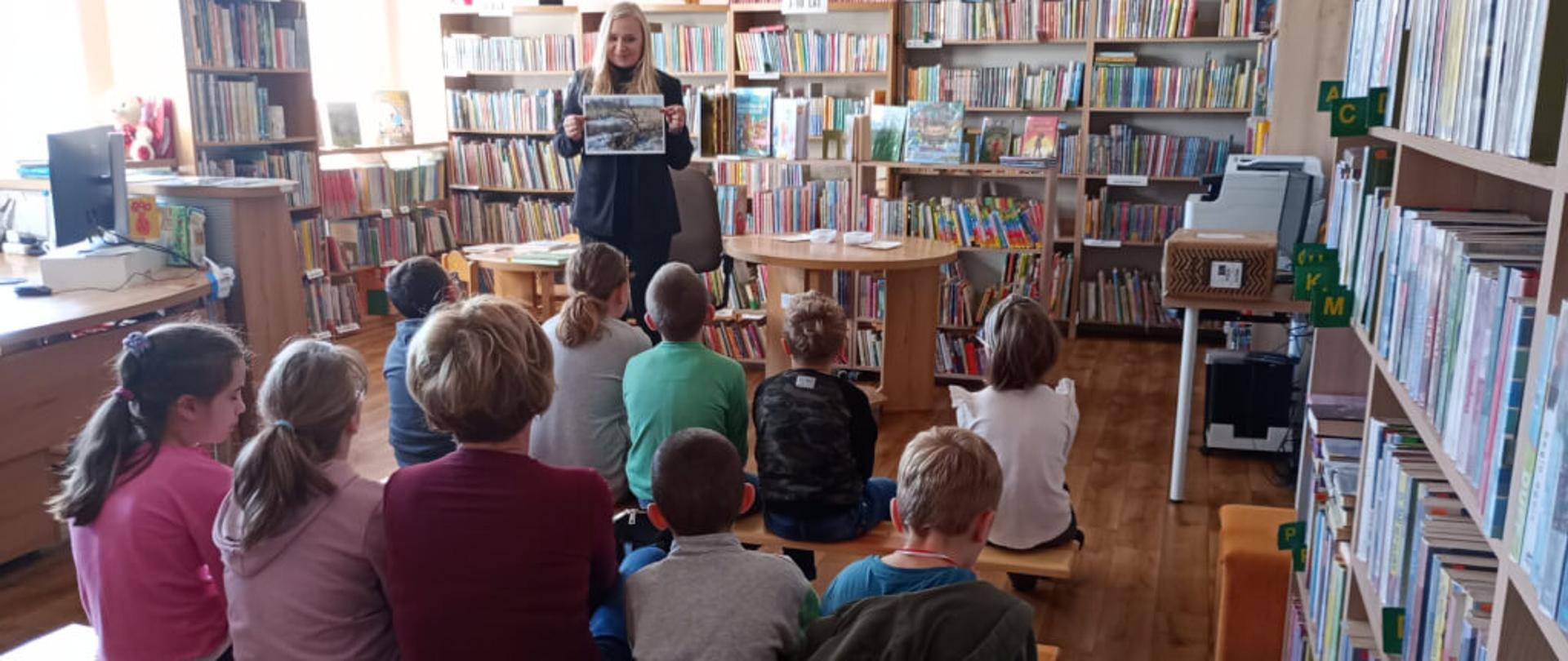 Zdjęcie przedstawia grupę dzieci siedzących na ławkach w bibliotece. Dzieci słuchają zagadek, które rozwiązują w ramach zajęć bibliotecznych