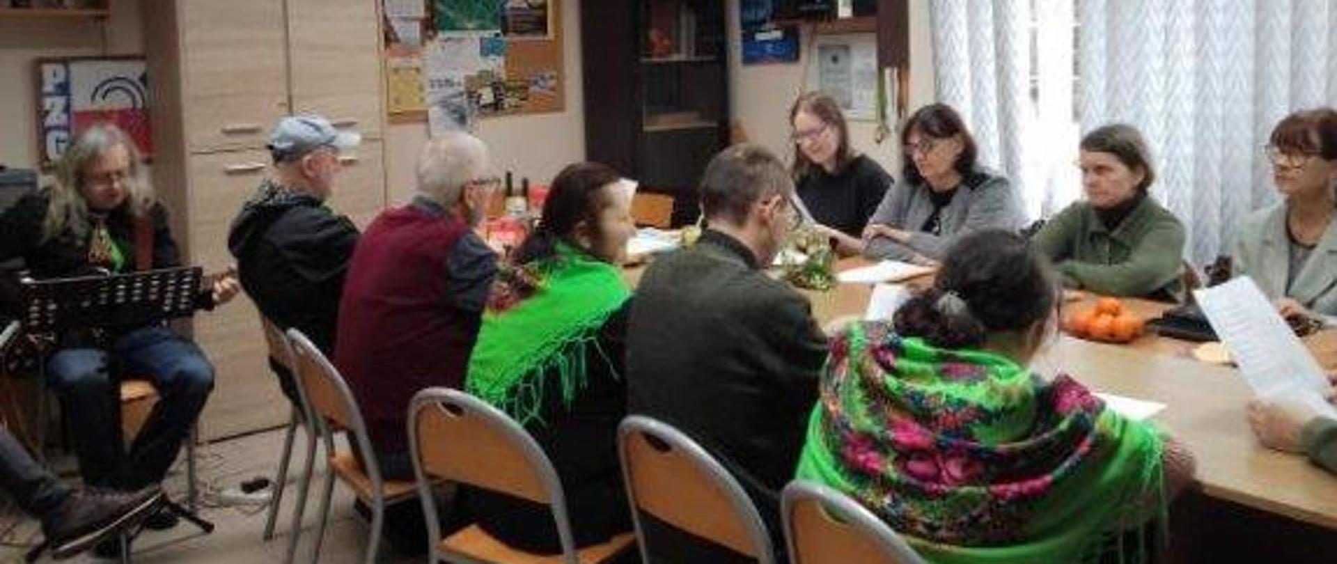 Uczestnicy spotkania siedzą przy stole, wspólnie kolędują