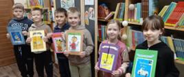 Uczniowie klasy 2 trzymają w ręku wykonane przez siebie Lapbooki czyli obrazkowe streszczenia przeczytanych książek, w tle regały biblioteczne