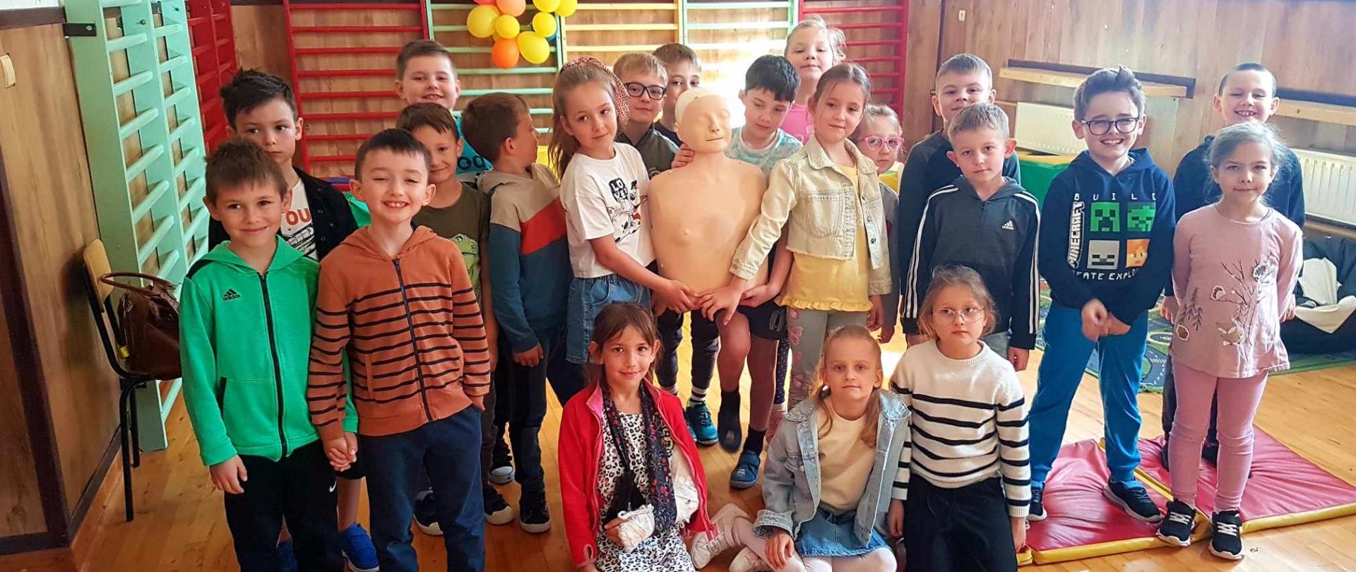 Uczniowie klasy pierwszej na sali świetlicy szkolnej wraz z fantomem uczestniczą w zajęciach z zakresu udzielania pierwszej pomocy przedmedycznej