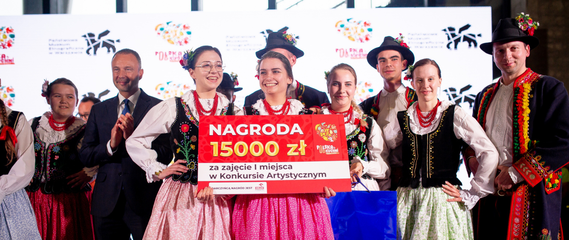 Zdjęcie promujące Festiwal Kół Gospodyń Wiejskich - Polska od Kuchni. Fotografia przedstawia zwycięzców w konkursie artystycznym, uczestnicy w strojach ludowych trzymają w rękach planszę z informacją o kategorii oraz wysokości nagrody, opiewającej na piętnaście tysięcy złotych.