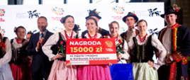 Zdjęcie promujące Festiwal Kół Gospodyń Wiejskich - Polska od Kuchni. Fotografia przedstawia zwycięzców w konkursie artystycznym, uczestnicy w strojach ludowych trzymają w rękach planszę z informacją o kategorii oraz wysokości nagrody, opiewającej na piętnaście tysięcy złotych.