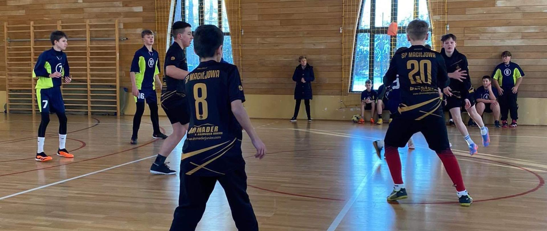 Na sali gimnastycznej Szkoły Podstawowej w Maciejowej grają w piłkę ręczną nasi zawodnicy w czarnych koszulkach ze złotymi napisami, widoczni są też zawodnicy drużyny przeciwnej w granatowo-zielonych koszulkach