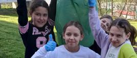 Cztery dziewczynki stoją przed szkołą, trzymają wspólnie uniesiony w górę zielony worek, na rękach mają niebieskie rękawice gumowe