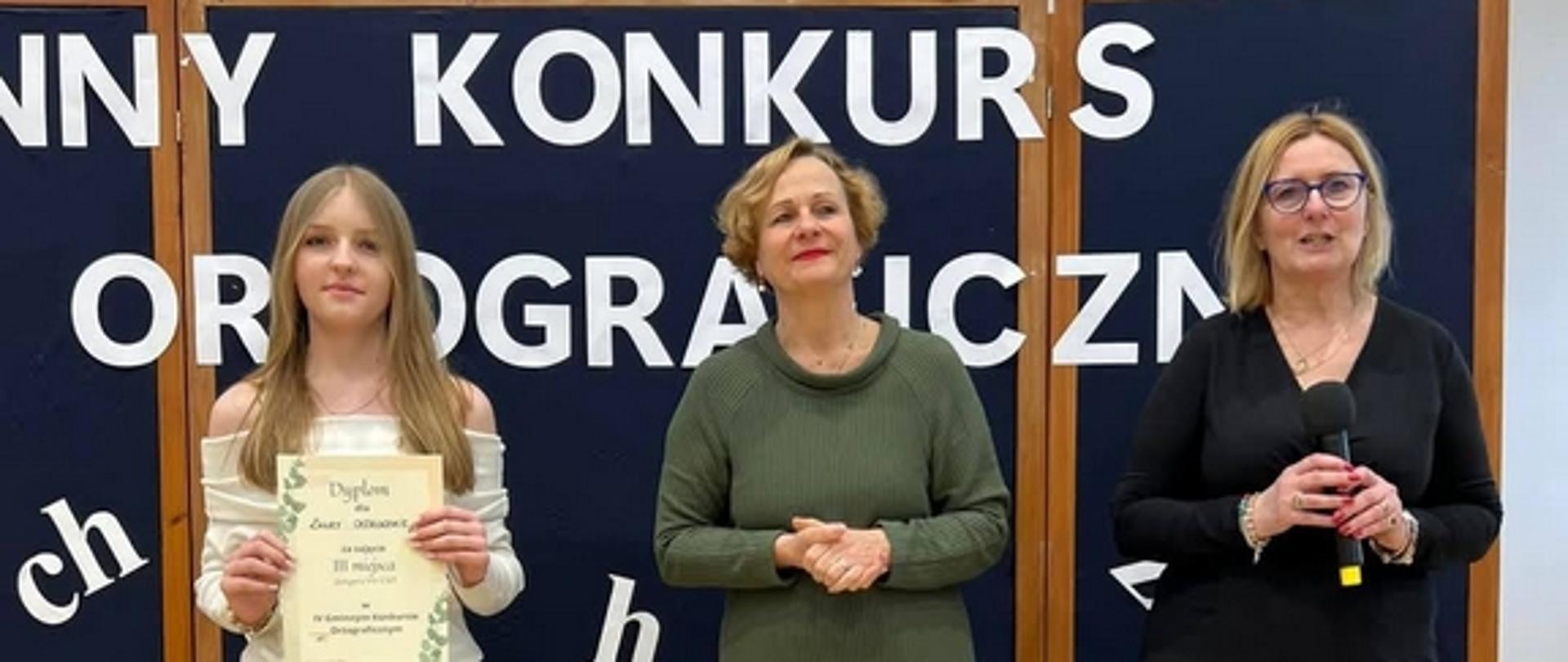 Na tle dekoracji z napisem Gminny Konkurs Ortograficzny stoją dwie kobiety, a obok nich dziewczynka z dyplomem w ręce.