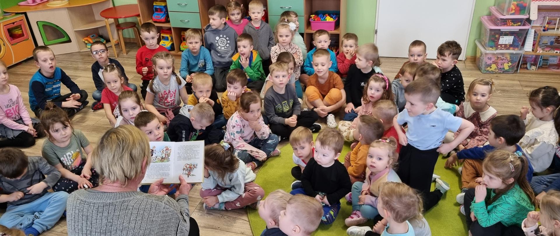 Na zdjęciu dzieci siedzą w sali i słuchają bajki czytanej przez bibliotekarza