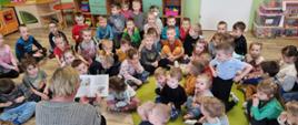 Na zdjęciu dzieci siedzą w sali przedszkolnej i słuchają bajki czytanej przez bibliotekarza