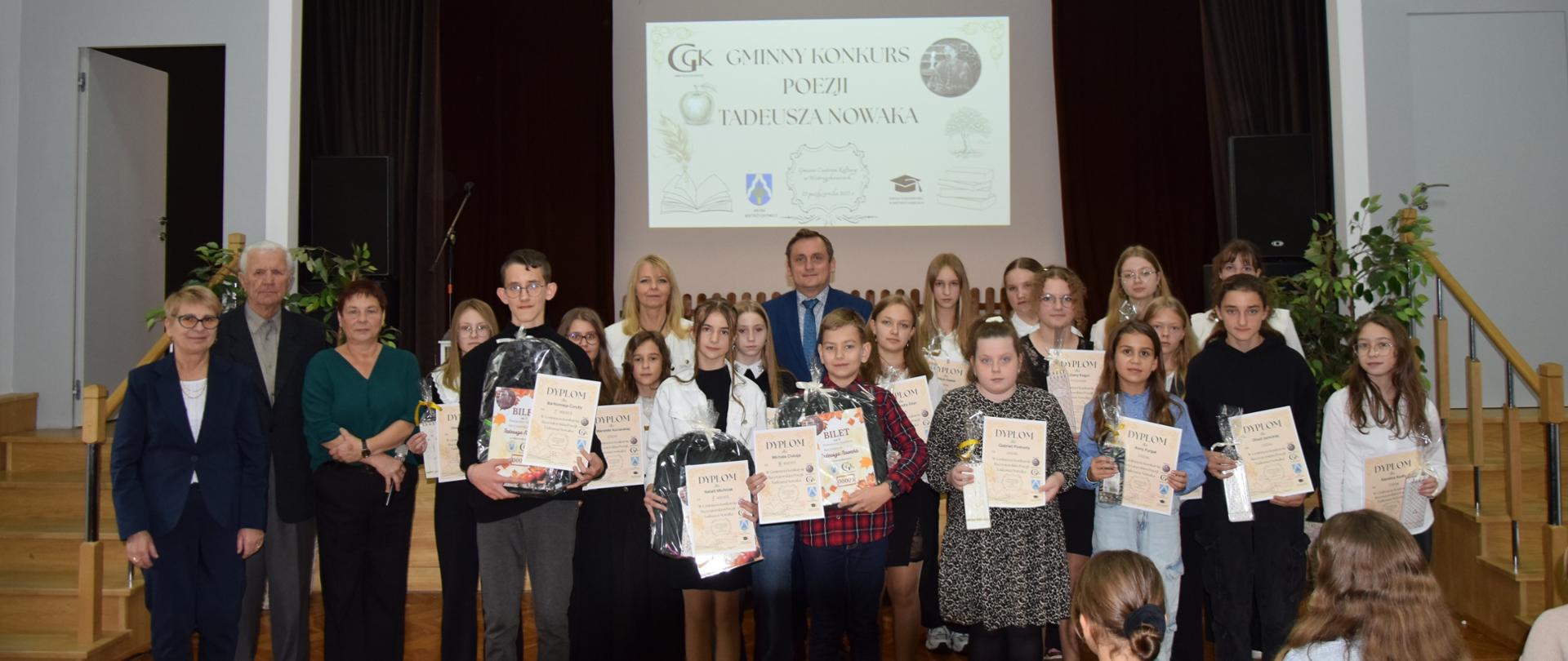 Grupa uczniów i nauczycieli pozuje na scenie z dyplomami i nagrodami podczas Gminnego Konkursu Poezji imienia Tadeusza Nowaka. W tle widoczny jest ekran z nazwą wydarzenia oraz dekoracje sceniczne.