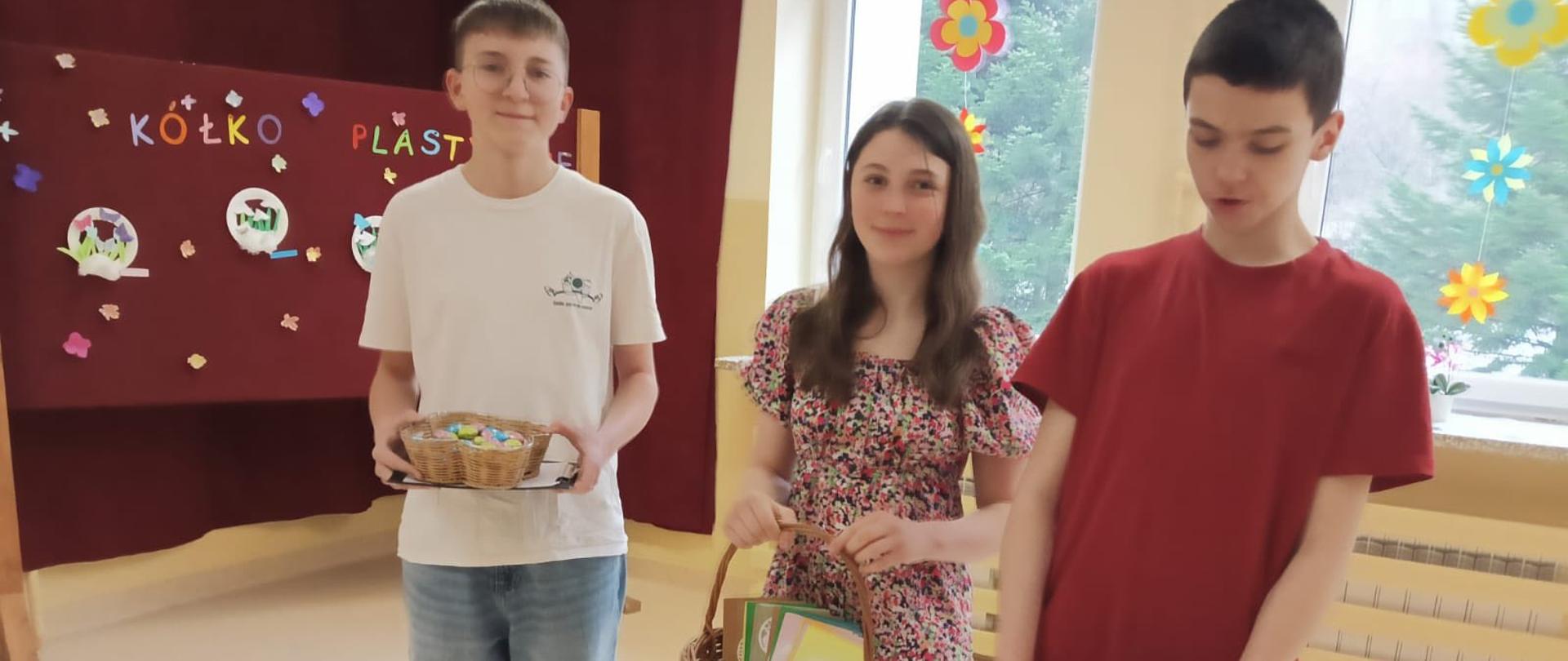 Przedstawiciele Samorządu Uczniowskiego w składzie 2 uczniów i 1 uczennica trzymają w ręku koszyki z czekoladowymi jajeczkami oraz wielkanocnymi kartkami