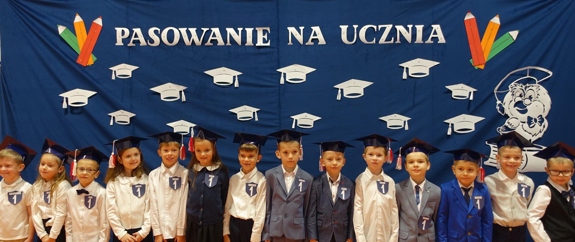 Pierwszoklasiści w biretach stoją na tle dekoracji - Pasowanie na ucznia