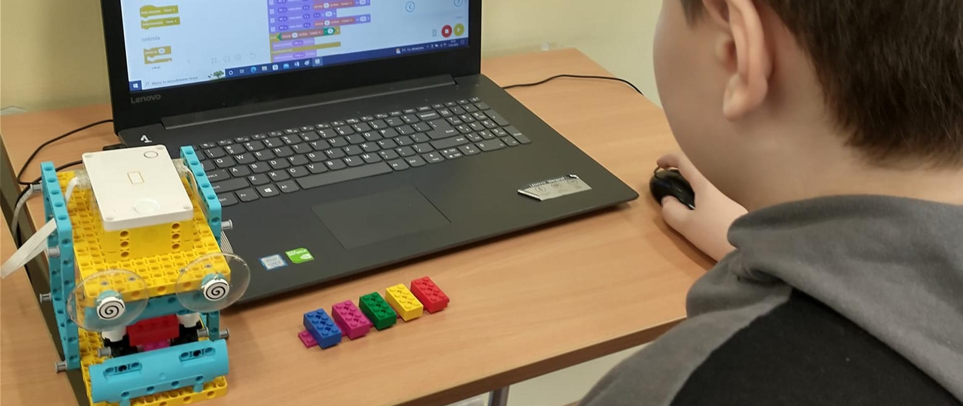 Uczeń siedzi przed laptopem i układa program uruchamiający zbudowanego robota. Po lewej stronie laptopa stoi robot zbudowany z kloców lego. Przed komputerem leżą klocki koloru niebieskiego, różowego, zielonego, żółtego i czerwonego.