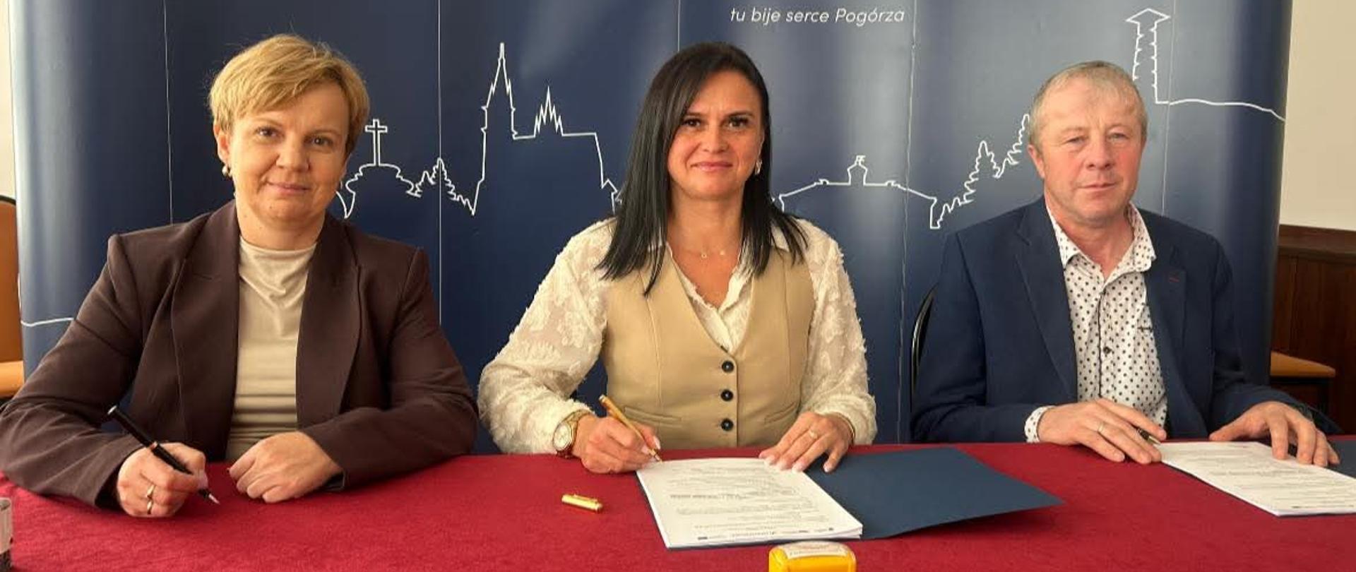 Trzy osoby siedzące na tle granatowej ściany z napisem gmina Ciężkowice