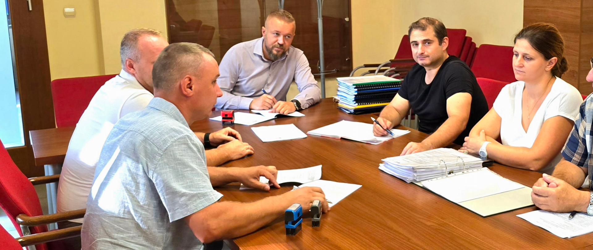 Kilka osób siedzi przy stole w jasno beżowej sali z elementami drewnianymi.
