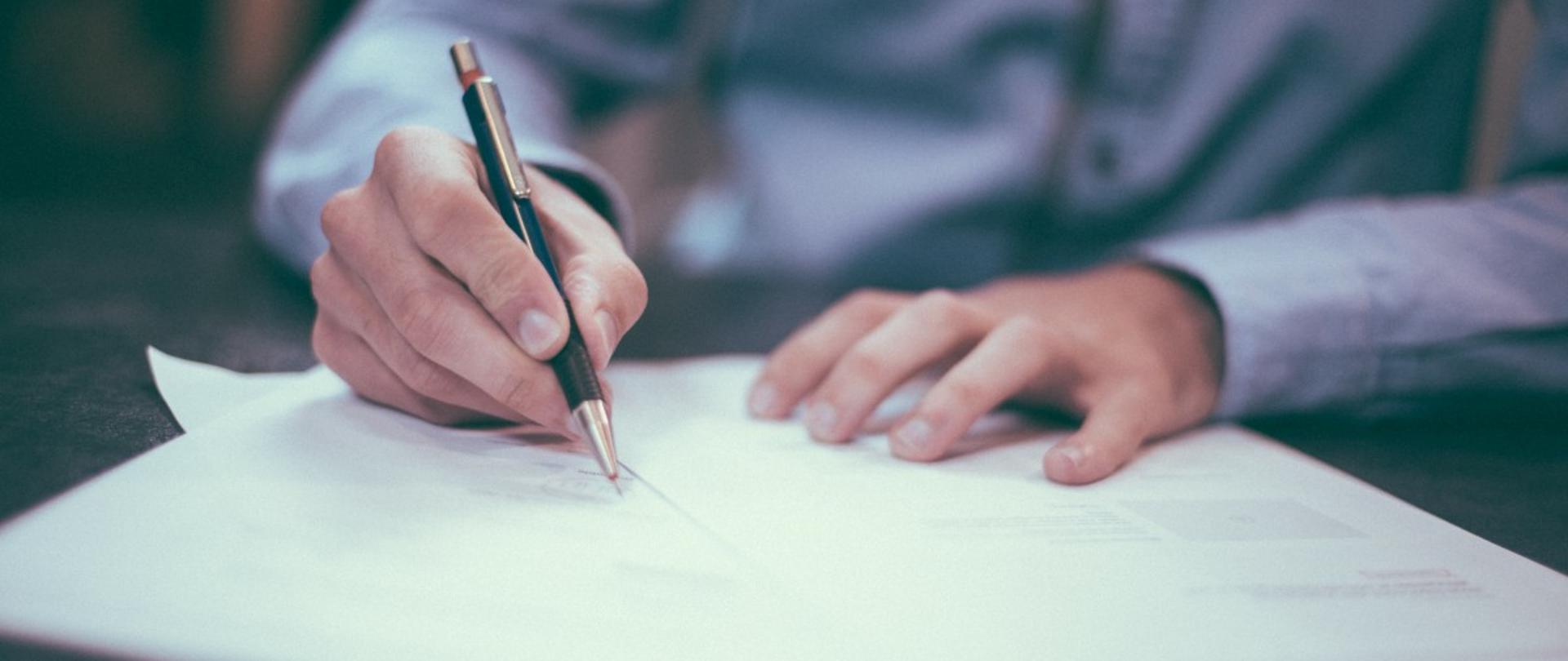 Na zdjęciu jedna osoba trzymają w ręce długopis i podpisująca dokumenty leżące na stole.