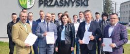 Zdjęcie przedstawia władze Powiatu Przasnyskiego oraz przedstawicieli wykonawcy, firmy Rembud po podpisaniu umowy na budowę basenów odkrytych. Od lewej stoją Wicestarosta Waldemar Trochimiuk, Starosta Przasnyski Krzysztof Bieńkowski, Skarbnik Beata Iwona Czaplicka, dwaj przedstawiciele firmy Rembud, w tle młodzież szkolna.